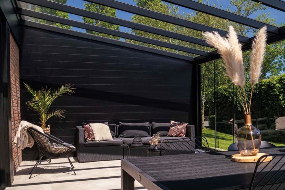 Moderner Pergola-Loungebereich mit schwarzer Aluminiumkonstruktion, Glasschiebeelementen und stilvoller Outdoor-Lounge im Gartenbereich.